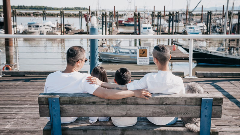 A same-sex couple and their children sit on a dock bench.